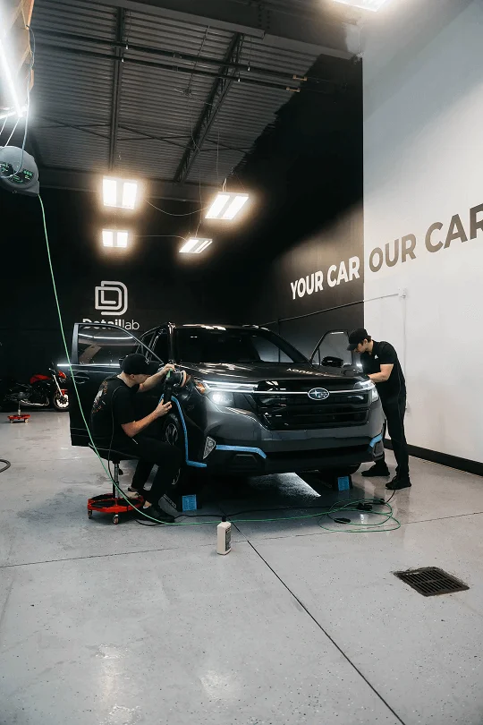 Detail Lab. Two professional detailers working on a dark-colored Subaru SUV inside a detailing shop. One detailer is polishing the front fender with a buffer while the other is cleaning the interior with the driver's door open. The shop's branding, DetailLab, and the text YOUR CAR OUR CARE are visible on the black and white walls.