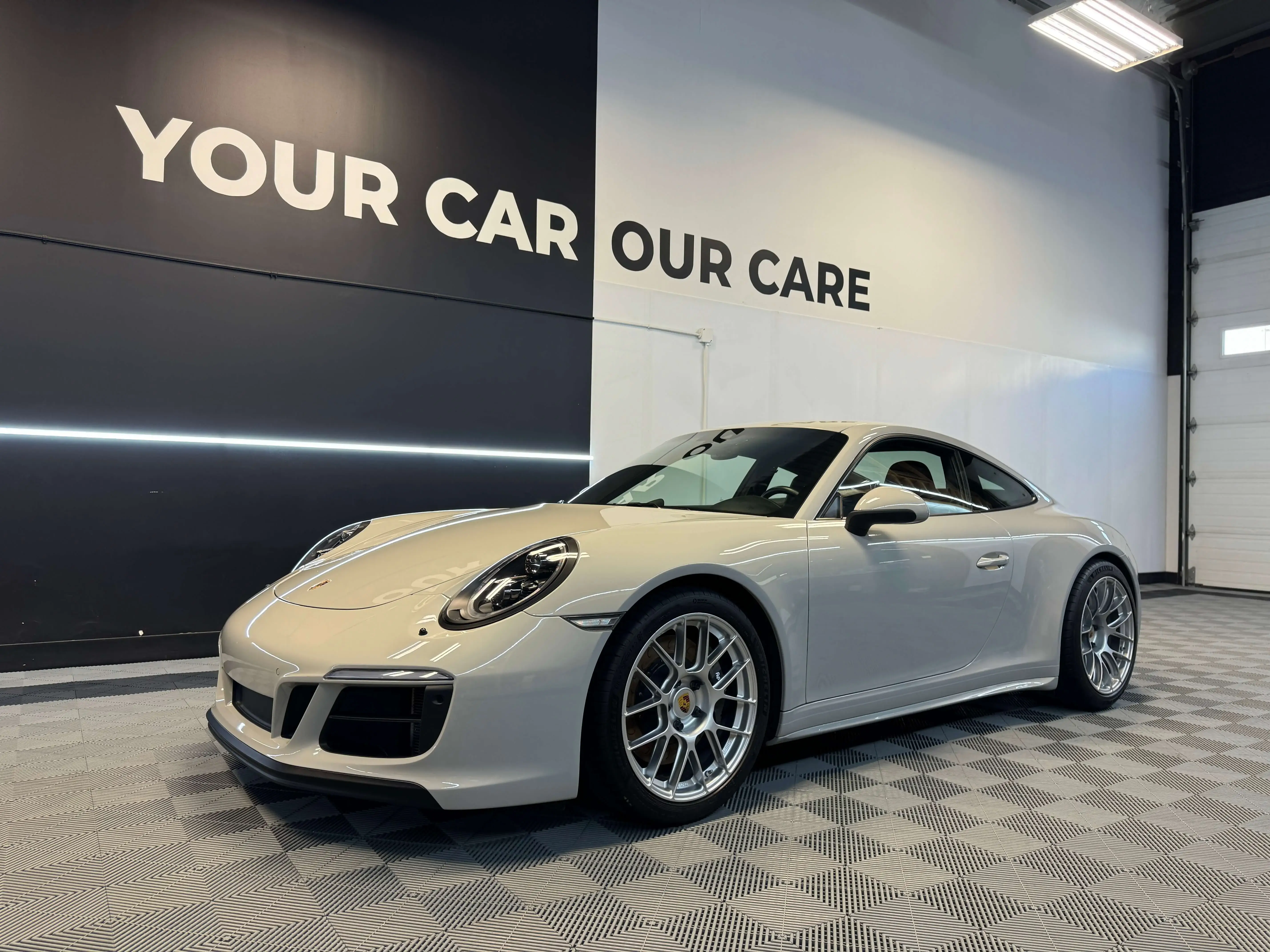 Detail Lab. White Porsche 911 coupe parked inside a detailing studio with a checkered floor. The polished exterior reflects the overhead lighting. The background wall displays YOUR CAR OUR CARE.
