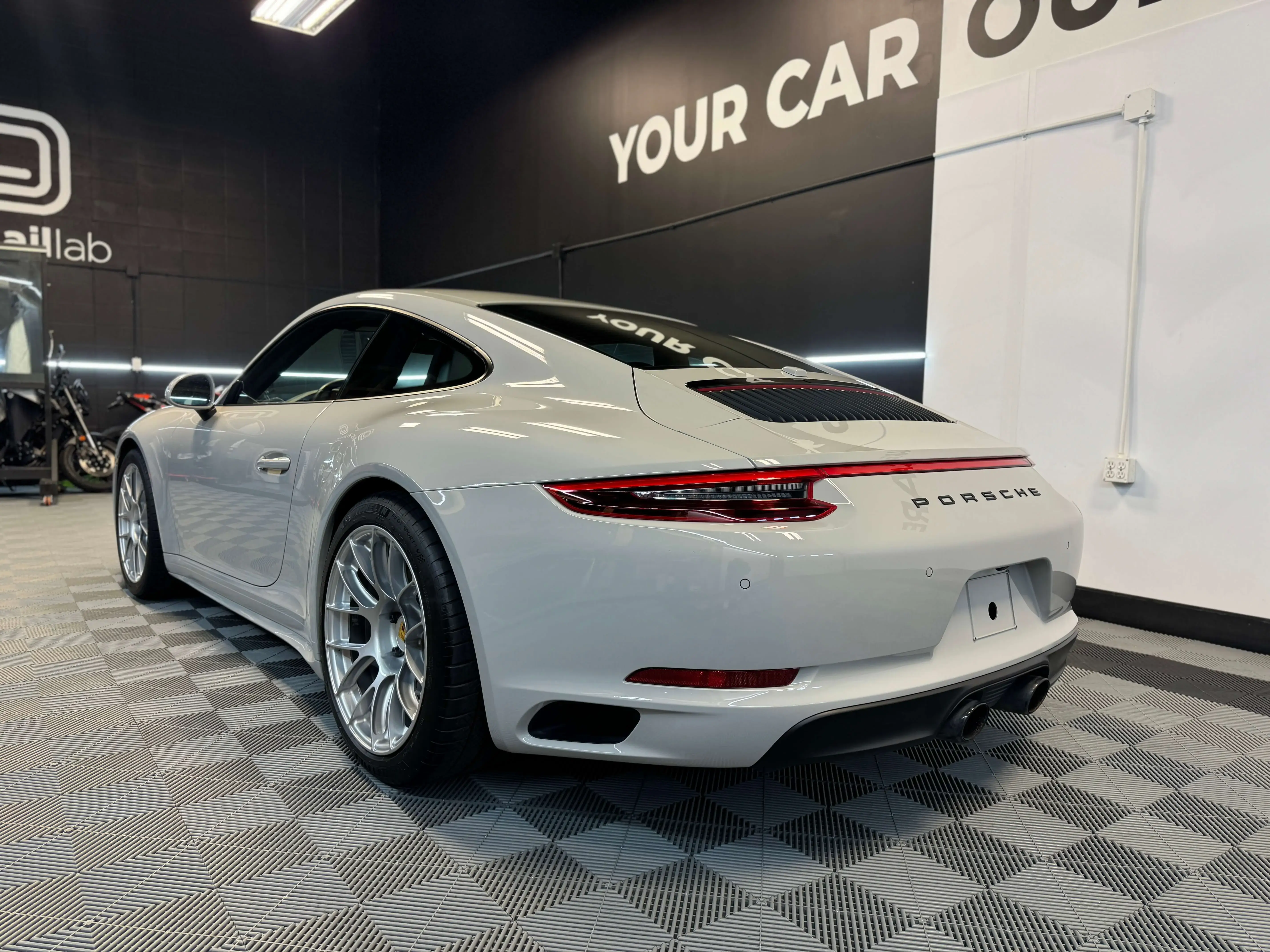 Detail Lab. Rear view of a white Porsche 911 in a detailing shop. The sleek design, rear spoiler, and Porsche branding are visible. The shop interior and motorcycles are reflected in the car’s glossy surface.