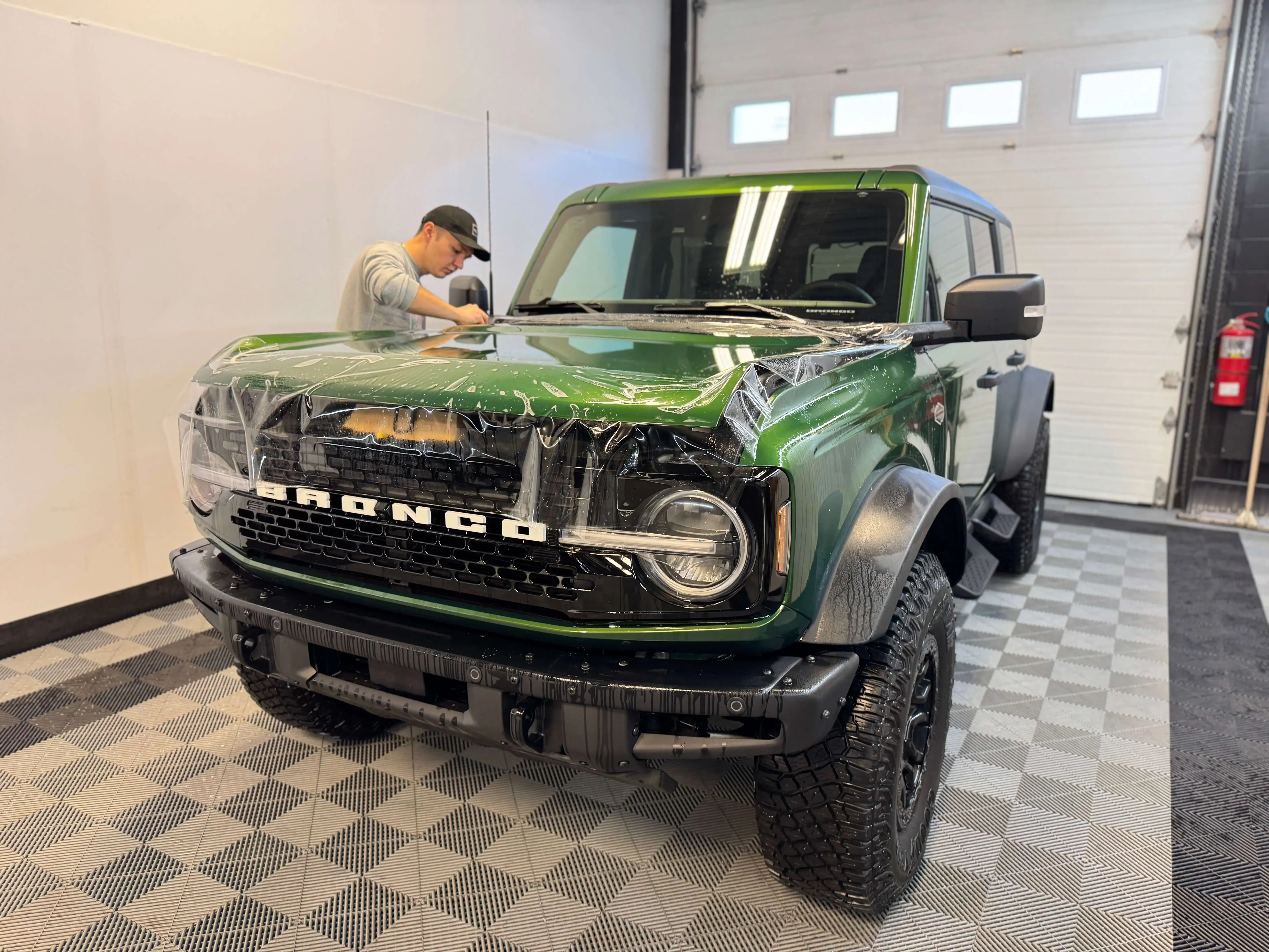 Detail Lab. Close-up of a technician applying paint protection film to the side of a green Ford Bronco Wildtrak. The clear film is being smoothed over the fender.