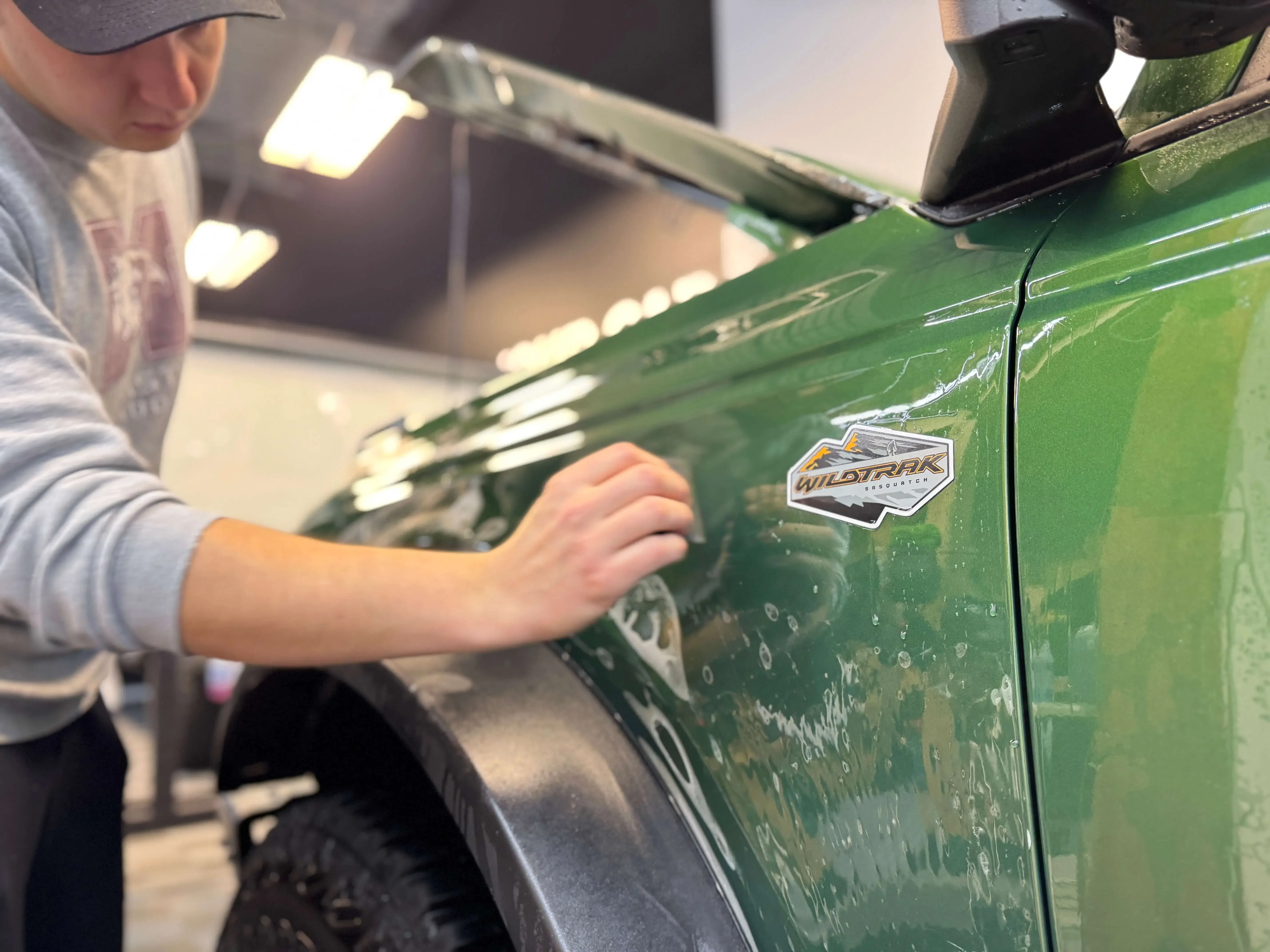 Detail Lab. Green Ford Bronco undergoing paint protection film installation. A technician is applying the film to the hood with water droplets visible on the surface. The garage door and equipment are in the background.