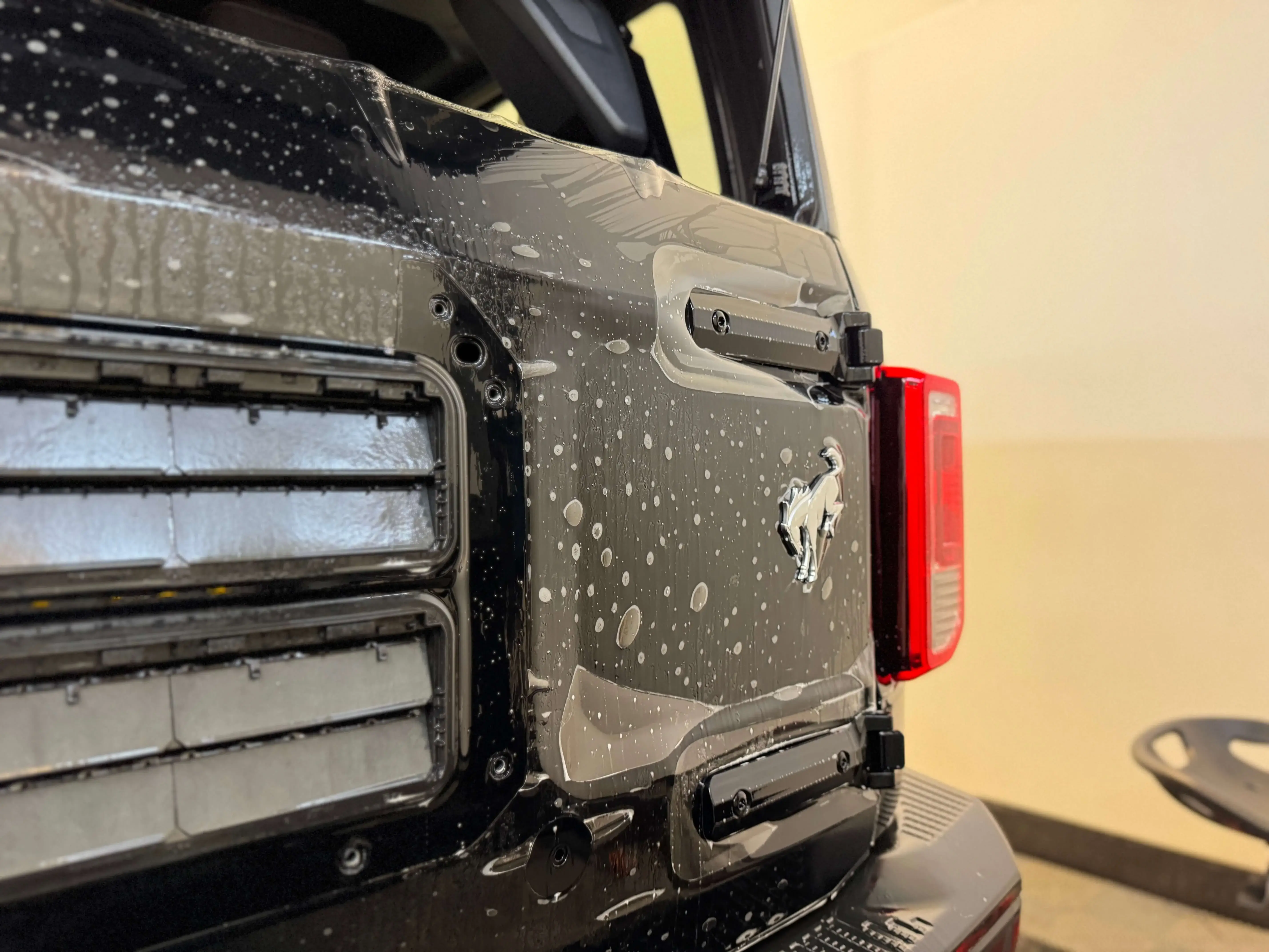 Detail Lab. Rear close-up of a black Ford Bronco with water droplets on the surface during paint protection film application. The taillight and silver Bronco emblem are visible.