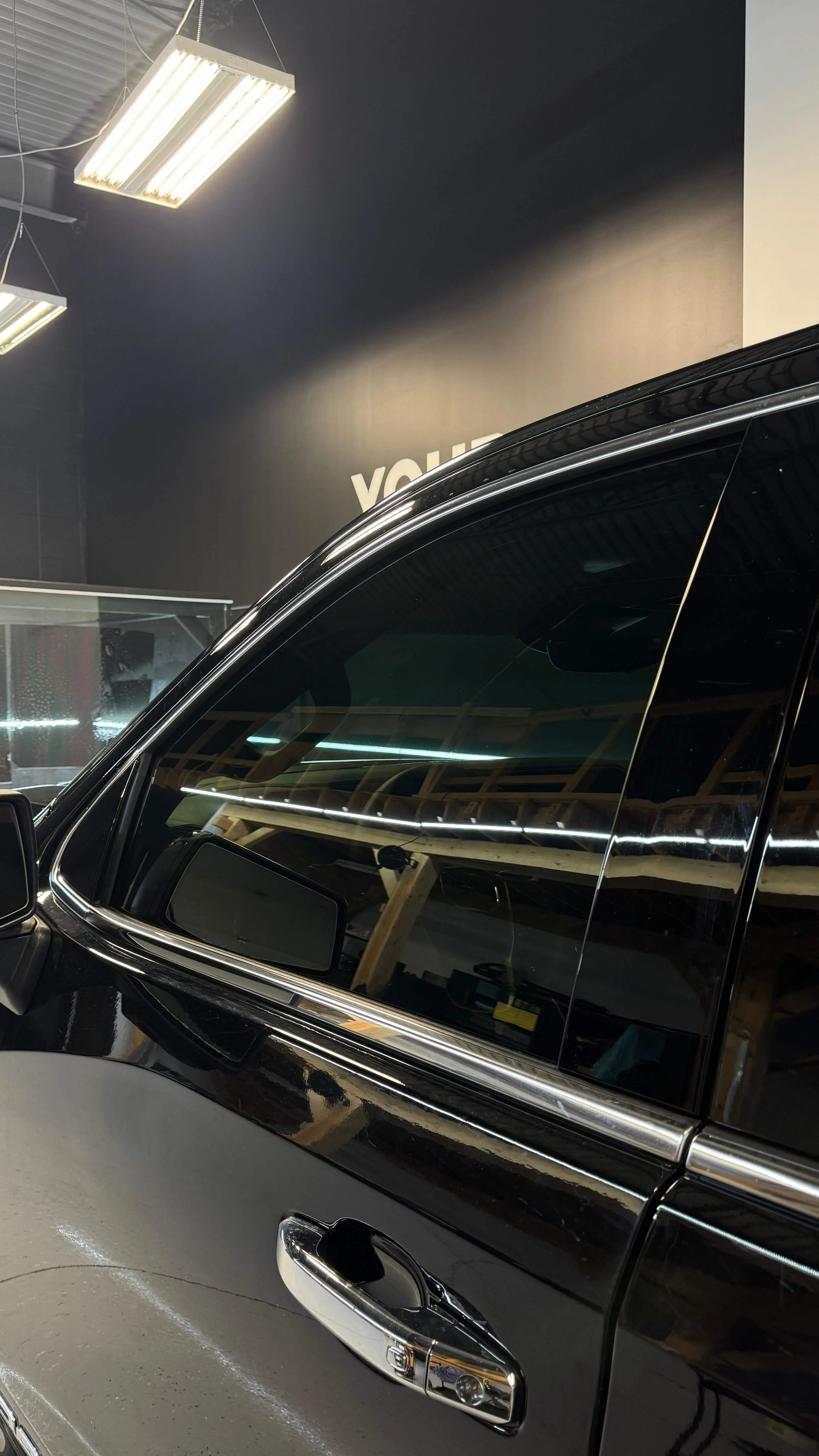Detail Lab. Close-up of a black truck’s tinted windows and chrome door handle. The reflections of overhead lights and shop interior are visible on the vehicle’s glossy surface.