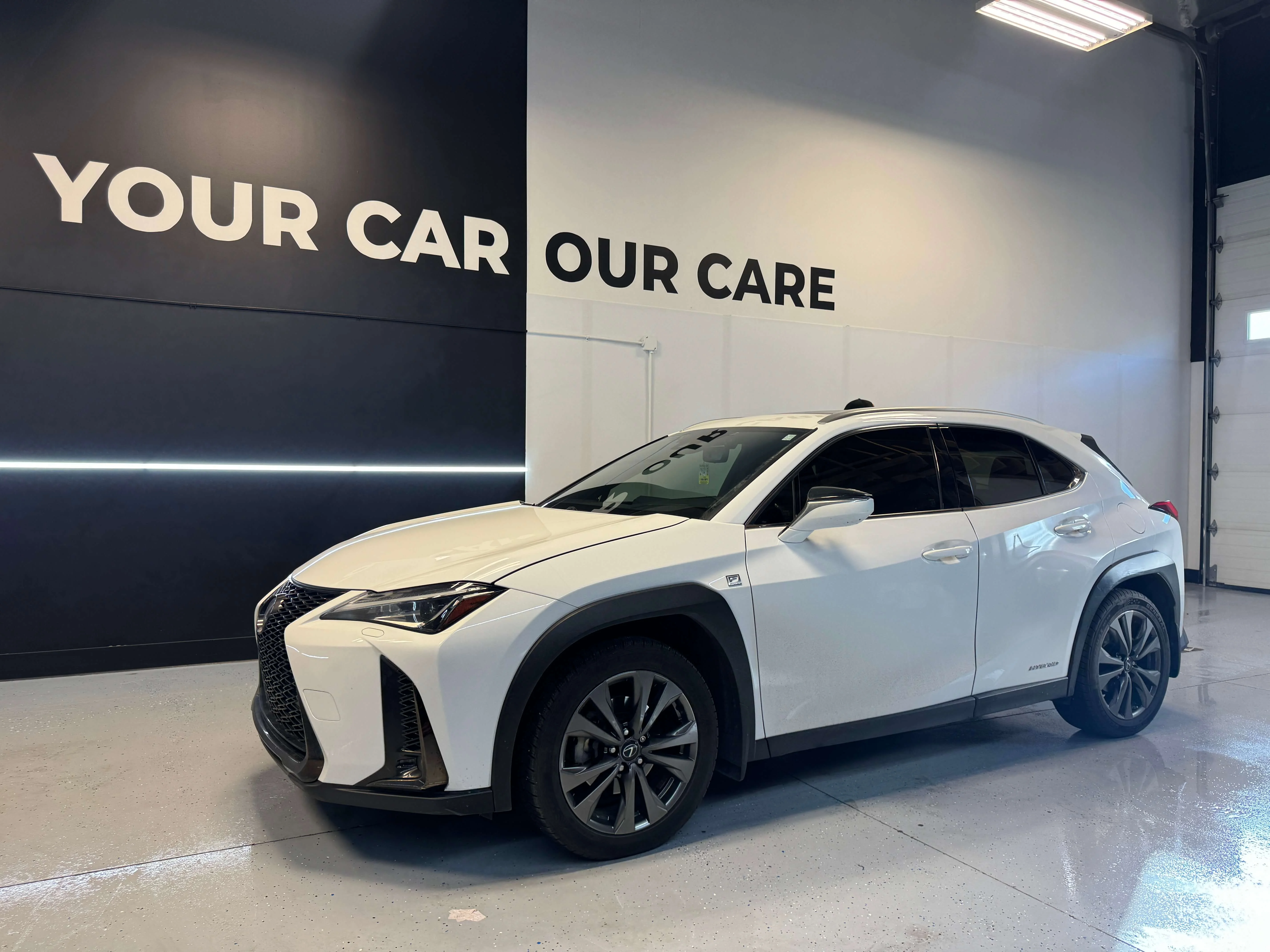 Detail Lab. White Lexus UX hybrid crossover with tinted windows parked inside a detailing studio. The wall behind the vehicle reads YOUR CAR OUR CARE.