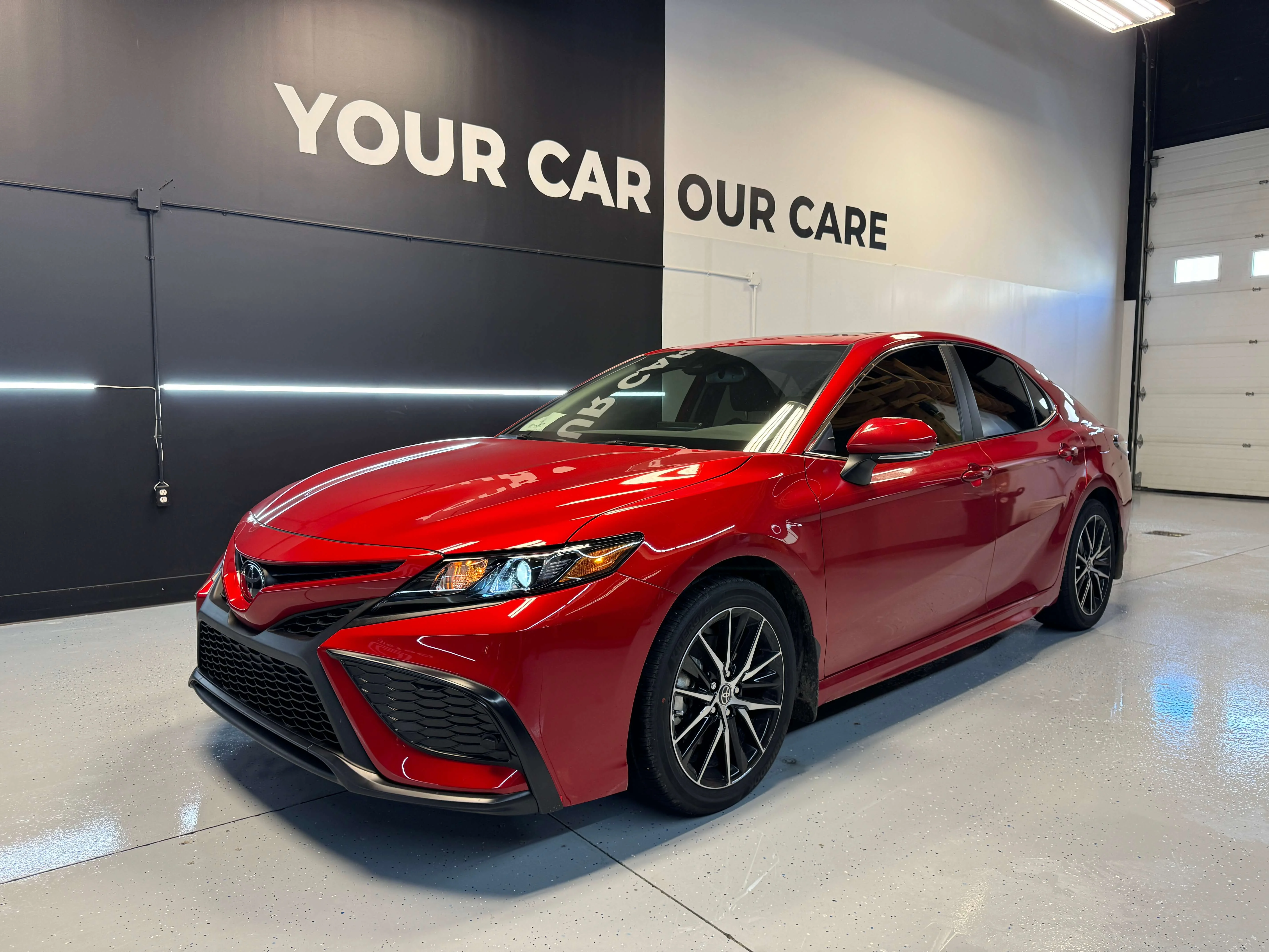 Detail Lab. Red Toyota Camry sedan with a glossy finish parked in a detailing shop. The bold red color contrasts with the black and white tones of the shop's interior. The words YOUR CAR OUR CARE are on the background wall.