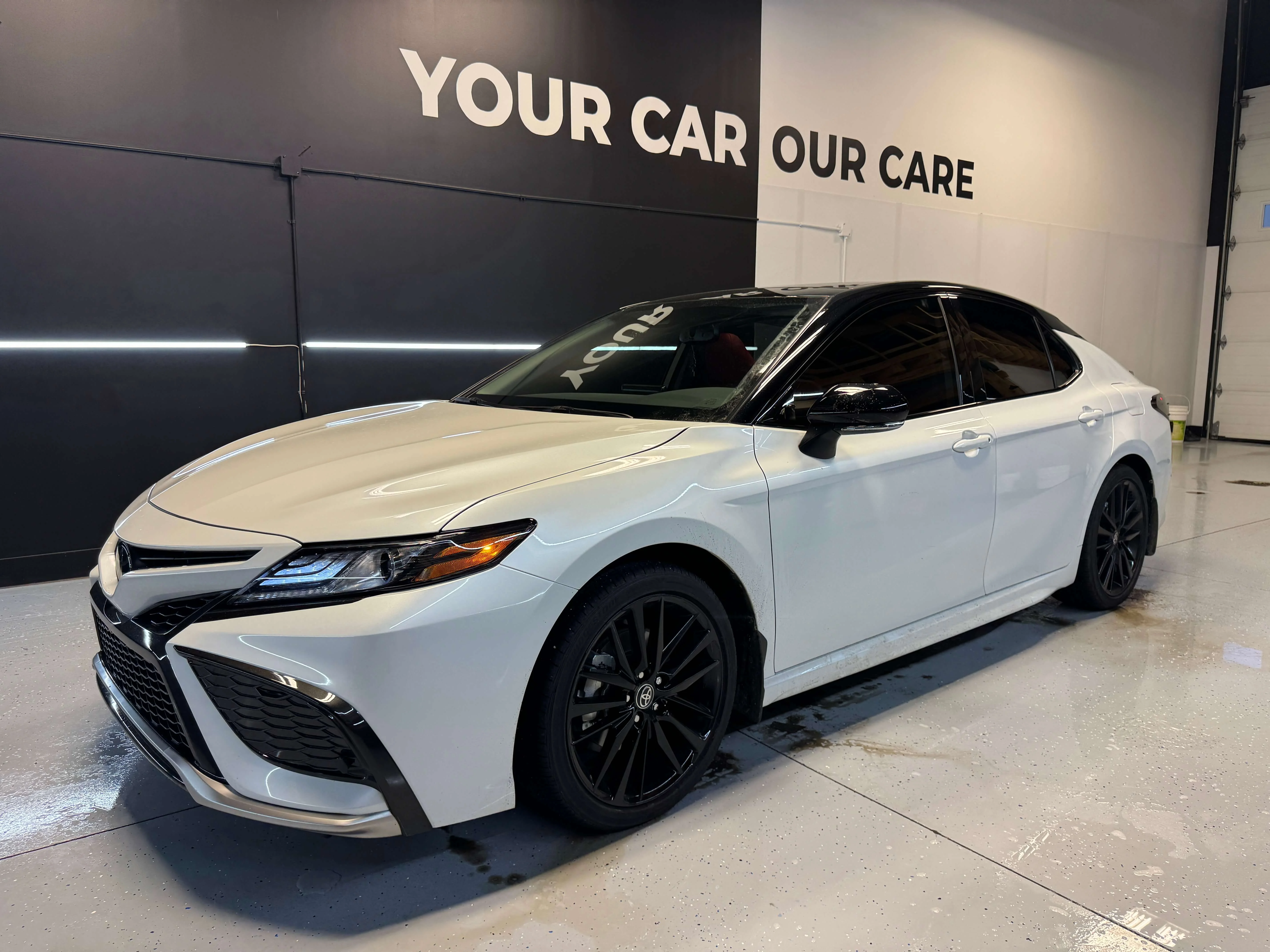 Detail Lab. White Toyota Camry with black rims and a glossy finish, parked inside a detailing shop under bright overhead lighting. The shop’s branding is visible in the background.