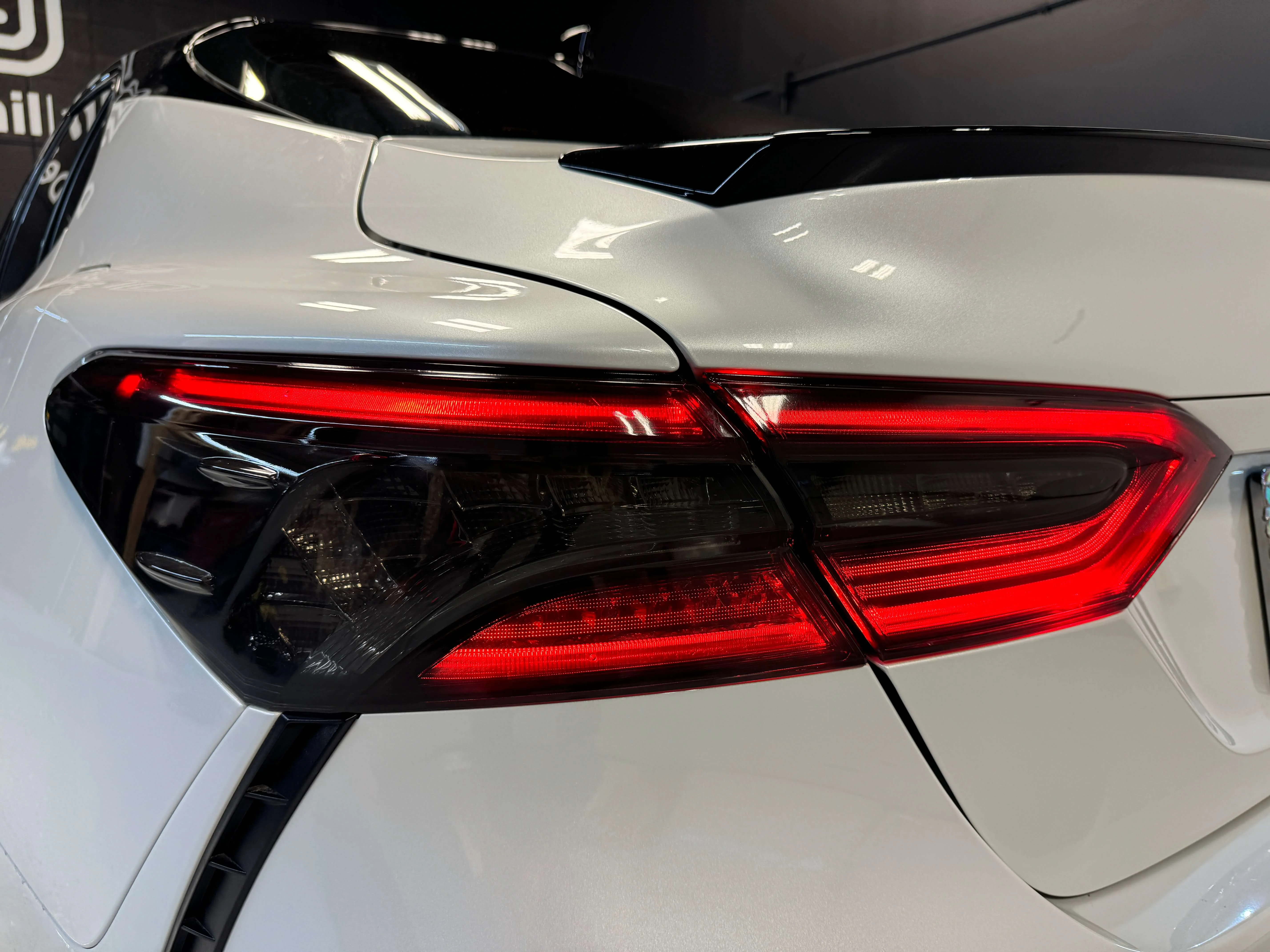 Detail Lab. Close-up of a white sedan’s rear taillight with a red and black design, featuring sharp lines and a glossy finish. The vehicle's spoiler and rear bumper are also visible.