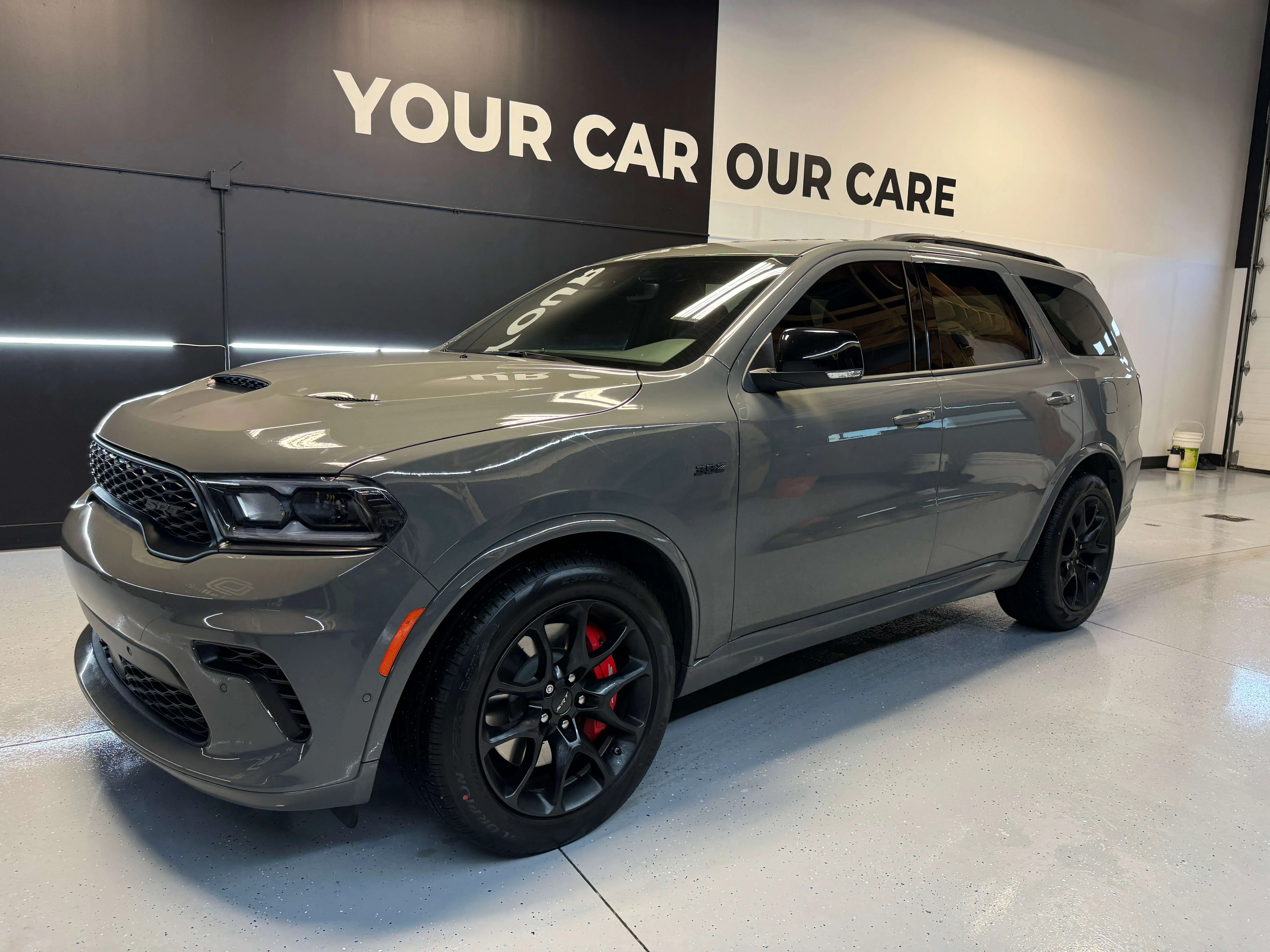 Detail Lab. Gray Dodge Durango SRT with black rims and red brake calipers, parked in a detailing shop under bright lighting. The polished body reflects the shop’s branding on the wall behind.